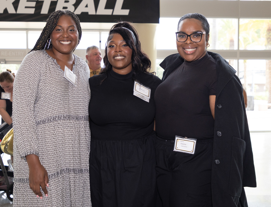 Three smiling women pose together at a basketball event, wearing name badges and dark outfits in front of a 'BASKETBALL' banner.