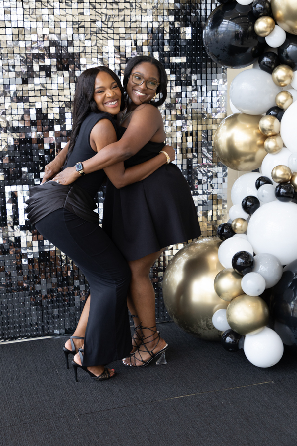 Two women in black dresses hug and smile in front of a silver sequin backdrop with a black, white, and gold balloon arch behind them.