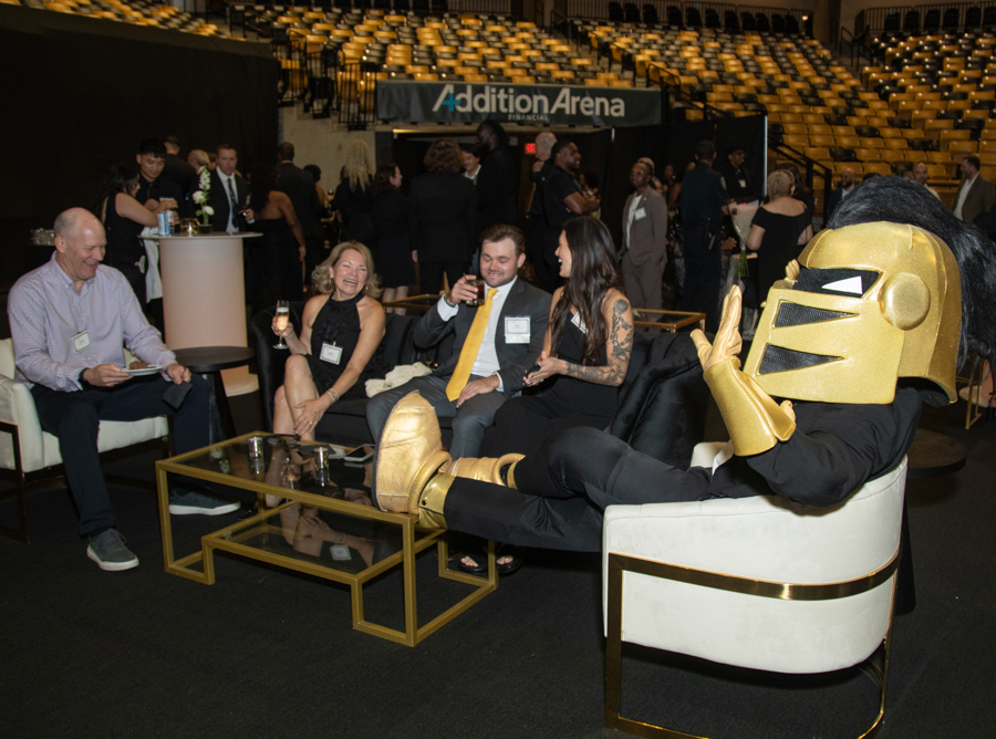 Group of professionally dressed attendees chatting and holding drinks on a lounge sofa at a reception, with a gold-helmeted mascot lounging nearby.