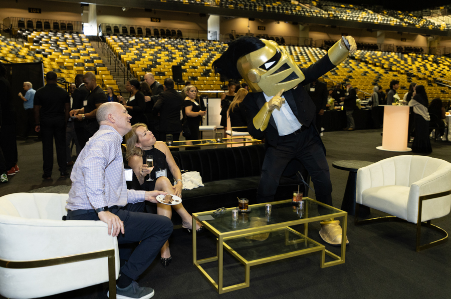 Man in a tuxedo wearing a large gold helmet mascot poses and leans toward a seating area of white couches and a gold-framed glass table as guests drink and chat at a formal event in a sports arena with yellow seats.
