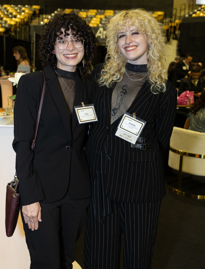 Two smiling women in black business attire with name badges posing at a formal reception.