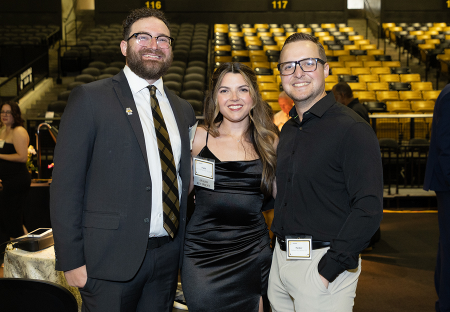 Three smiling adults in formal attire pose together at an event in a stadium arena, with name badges visible on their outfits.