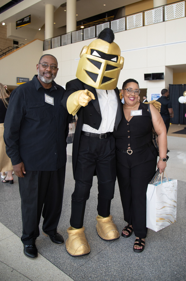 Three people pose for a photo with a gold knight mascot in a suit at a convention center. Name tags visible on two adults.