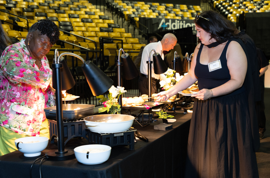 Woman in a black dress serves herself at a buffet line with chafing dishes and flowers in the center of a catering setup in a stadium venue of yellow seats.
