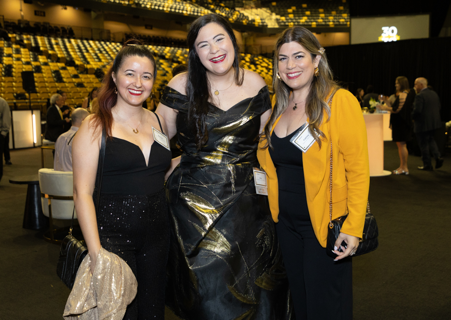 Three women in formal outfits pose together at an event, smiling with name badges visible on their chests nearby a stage area in the background, venue lights above.