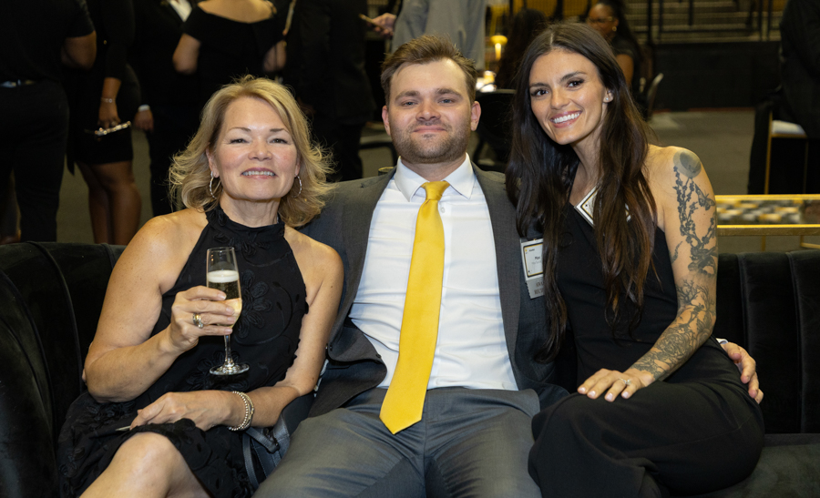 Three people pose on a black couch at a formal event: woman with champagne glass, man in a gray suit and yellow tie, and woman with a tattooed arm on the right.