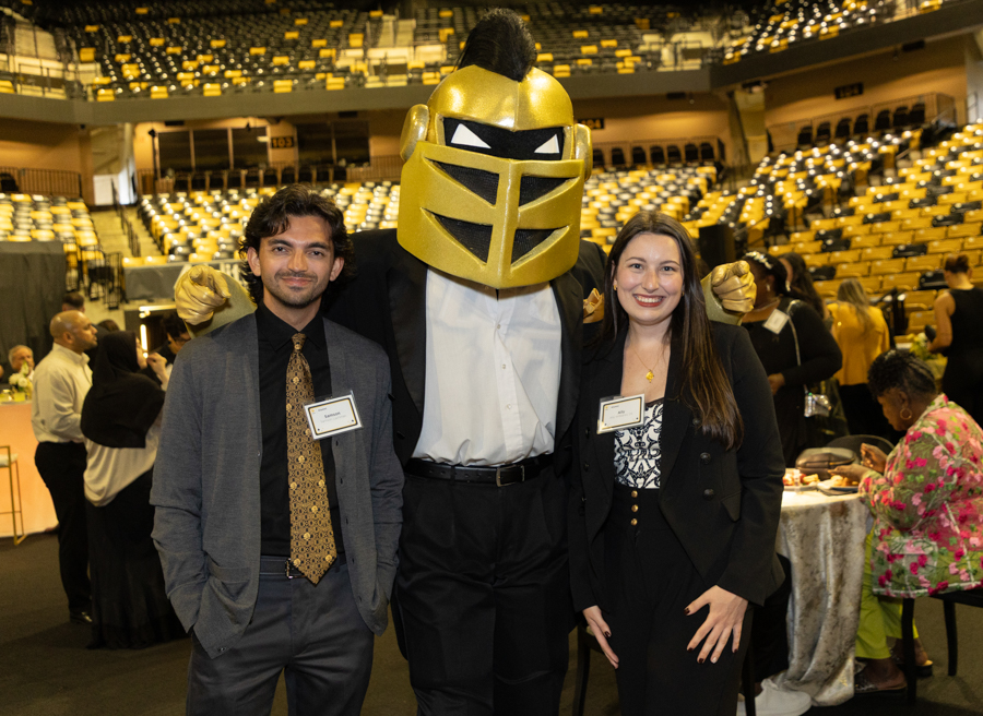 Two attendees pose with a large gold knight mascot at a formal event in a stadium arena, name badges visible on their jackets.