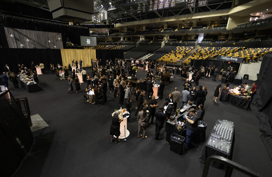 Large reception in an arena with guests mingling around high-top and round tables, buffet stations, and a stage backdrop far left.