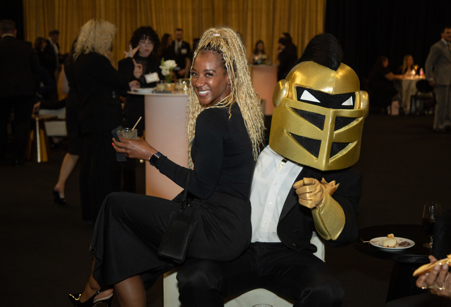 Smiling woman in a black dress sits with a person wearing a large gold knight helmet at a formal event, holding a drink.