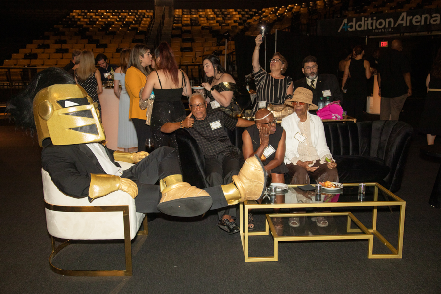 Group of guests at a formal event lounging on a sofa, including a person in a large gold helmet costume while others mingle nearby in an arena setting.
