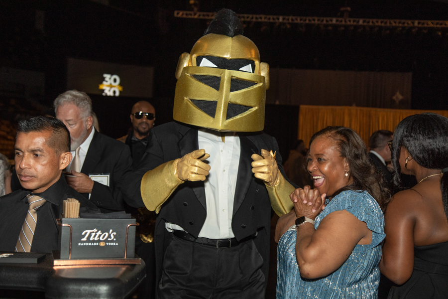 Gold knight helmet mascot in a tuxedo standing with guests at a formal event, a woman in a blue dress smiles beside him.
