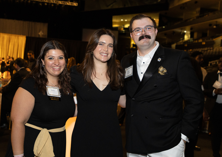 Three adults posing together at a formal event, two women in black dresses and a man in a dark suit with glasses and a mustache, smiling at the camera.