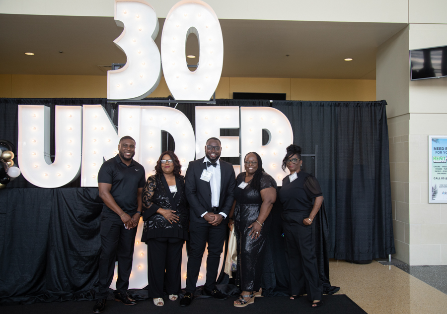 Five adults in formal wear pose in front of a large illuminated '30 Under' sign at an event staged indoors.