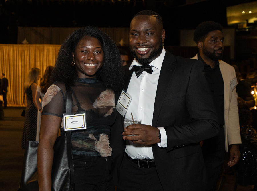 Two smiling attendees at a formal event posing for a photo; a woman in a sheer black top with a name tag stands beside a man in a black tuxedo and bow tie who is holding a drink.