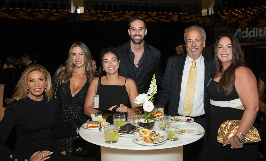 Six people in formal attire posing around a tall round table with food and drinks at a social event, floral centerpiece visible.