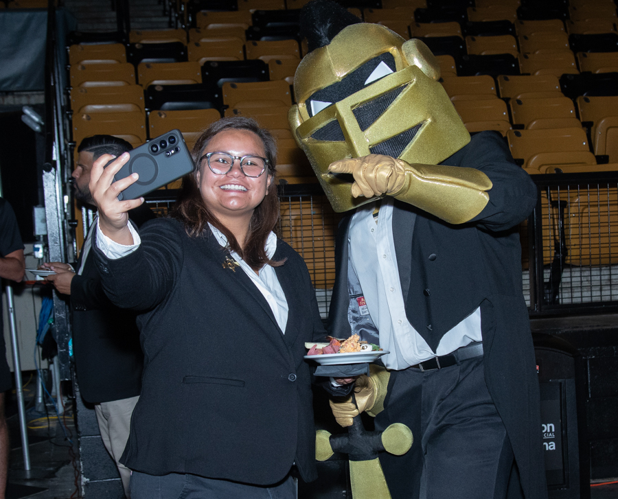 Woman in a blazer taking a selfie with a gold armored knight mascot, holding a plate of food, in a venue with yellow and black seats behind them.