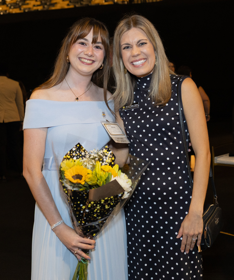 Two smiling women posing together at a formal event; the left woman wears a light blue off‑the‑shoulder dress and holds a bouquet of sunflowers, while the right woman wears a navy polka‑dot dress with a small purse.