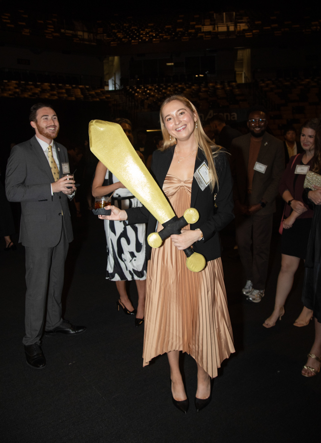 Smiling woman in a peach pleated dress and black blazer holds a large gold foam sword at a formal event with guests in the background.