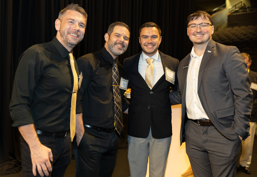 Four men in suits posing together at a formal event, smiling for a group photo with name tags visible on their jackets.