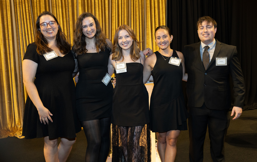 Five people in black formal wear pose together on a stage with gold curtains, smiling for a group photo at an event.