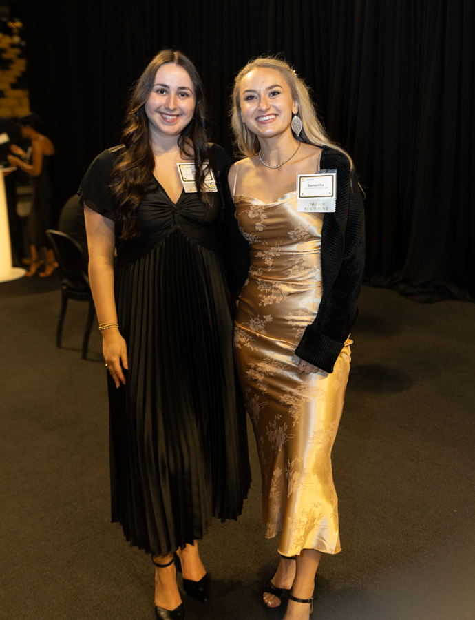 Two smiling women posing at a formal event: one in a black pleated dress, the other in a gold satin floral dress with a black cardigan, both wearing name badges.
