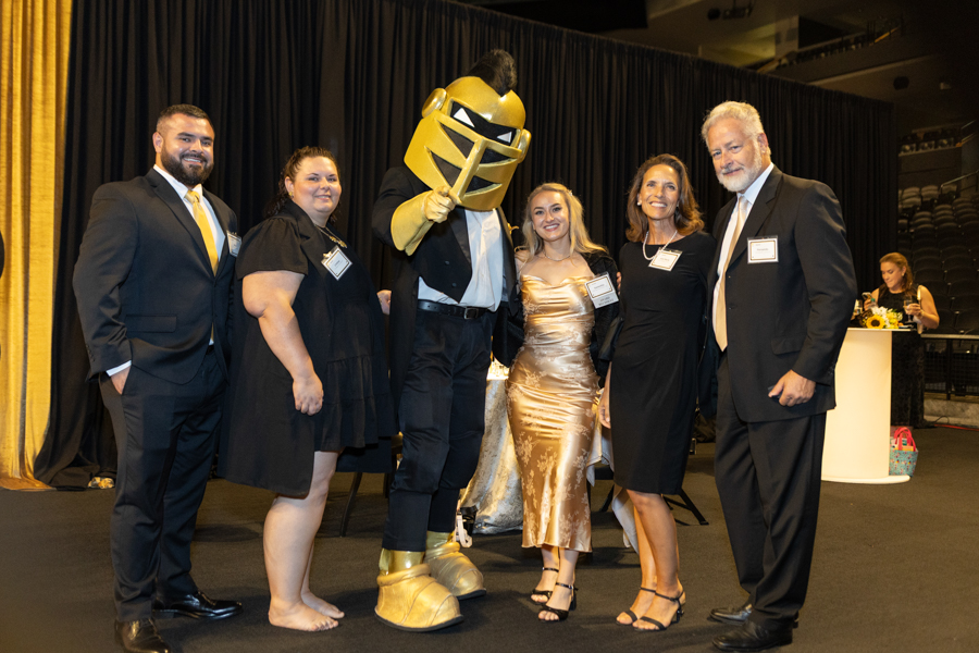 Group of six adults in formal attire posing with a costumed knight on a stage with black curtains in the background.