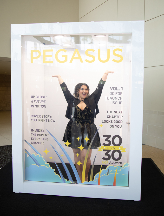 Woman in a black dress stands inside a large PEGASUS magazine cover display, arms raised, with colorful waves at the bottom and celebratory graphics.