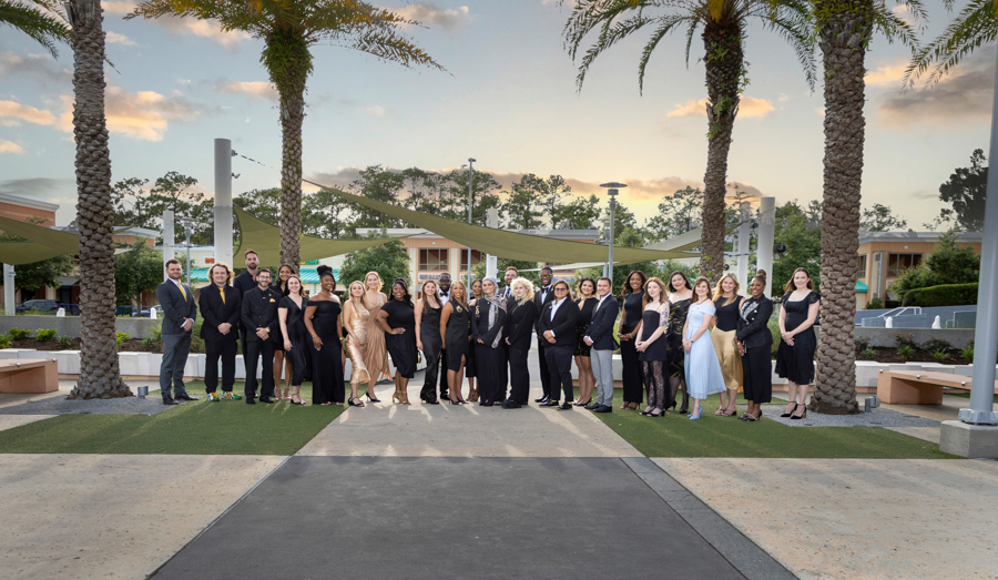 Group of about 30 people in formal attire posing outdoors under palm trees at sunset.