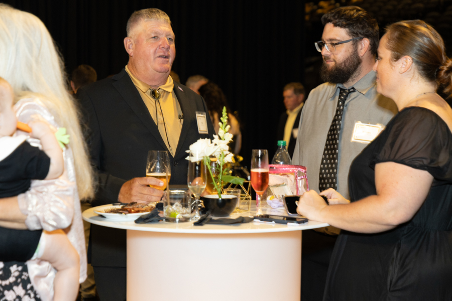 Man in a tan shirt and black blazer speaks to two guests at a standing round table with drinks and a small flower centerpiece at a formal gathering.