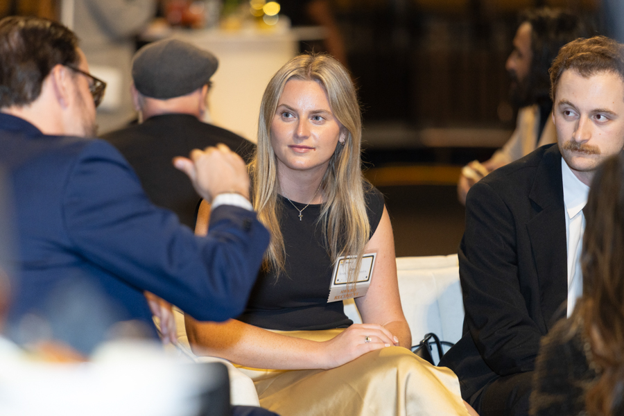 Blonde woman in a black top and beige skirt sits at a table with a name badge, listening to a man in a blue suit on the left.