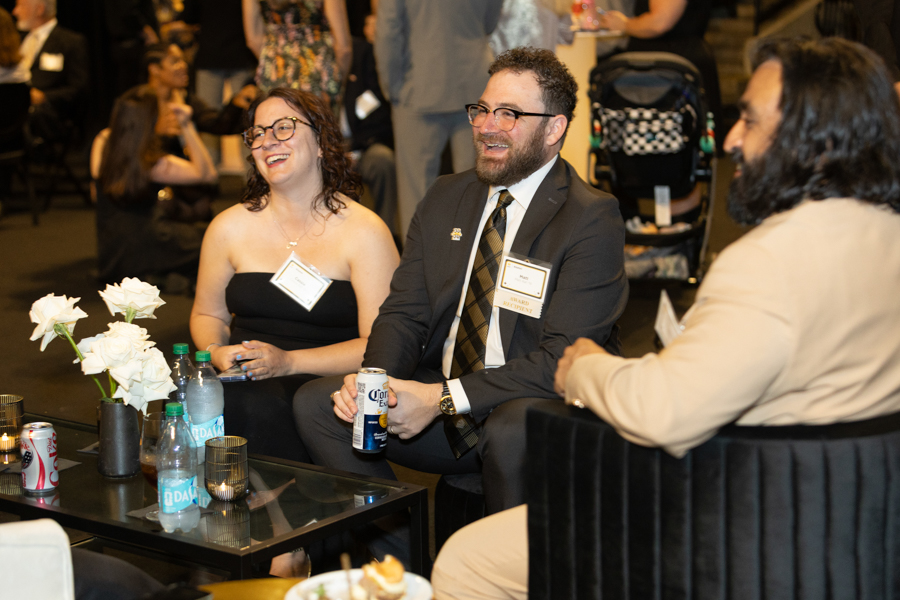 Two smiling adults sit together at a round table during a formal event, with name badges on their chests and drinks on the table near white flowers.