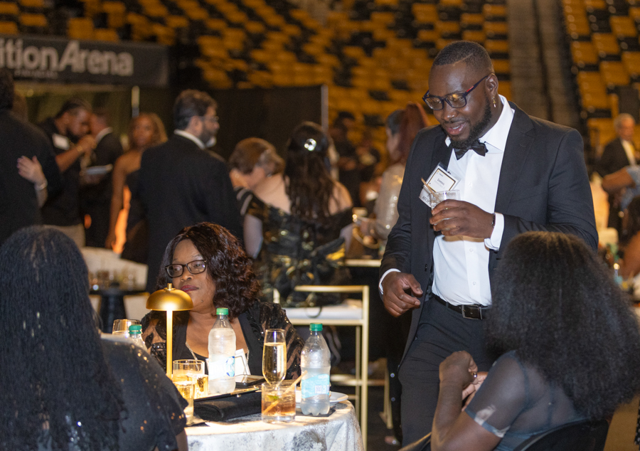 Man in a black suit with bow tie stands by a round banquet table, talking to seated guests at a formal event.