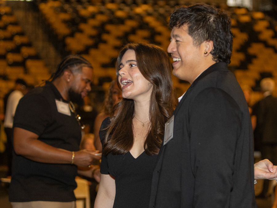 Two people are smiling and talking at a formal event, with a blurred audience in the background, wearing name badges.