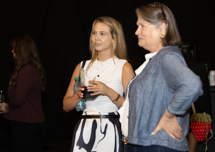 Two women at a social event; the woman in a white sleeveless top holds a drink while a second woman in a denim jacket stands beside her.