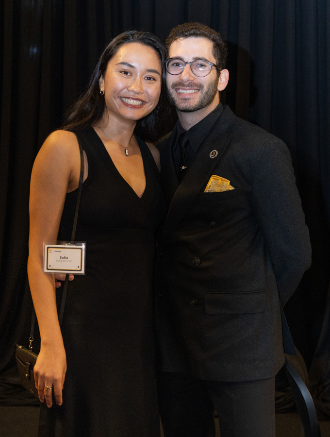 Couple in black formal wear posing together and smiling at a formal event, with a dark curtain backdrop.