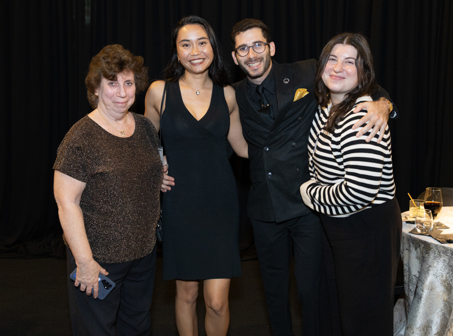 Group of four people posing together at a formal event, smiling at the camera. Woman on left wears a brown top, woman second from left in a black dress, man second from right in a black suit with a gold pocket square, woman on far right in a striped sweater and black pants. Audience table with drinks visible to the side.