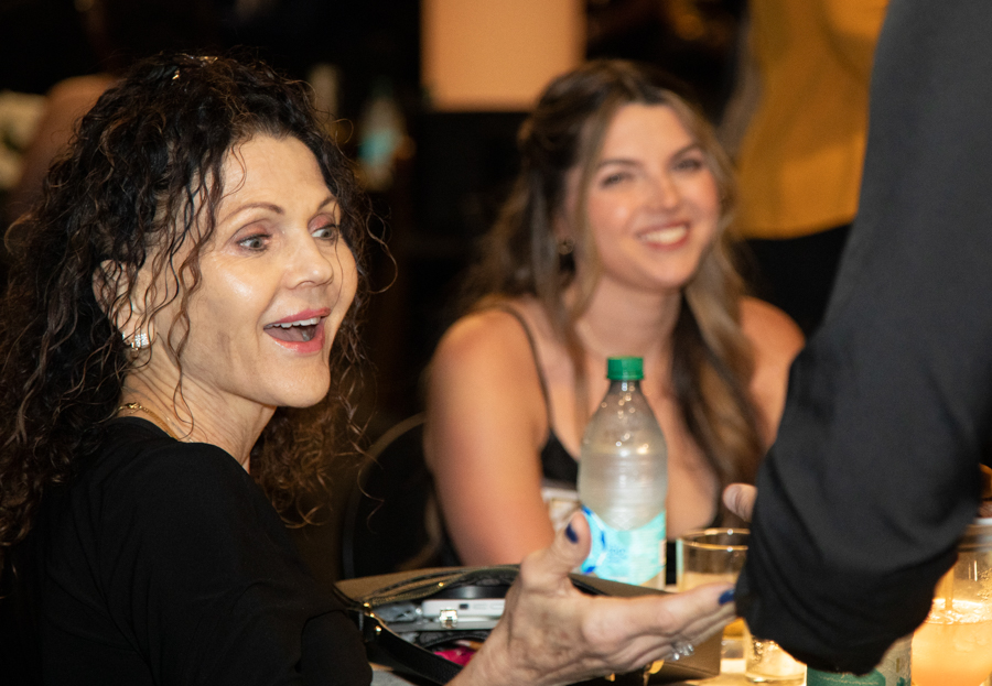Older woman with curly dark hair extends her hand in a greeting while seated at a table, a smiling woman sits in the background and a water bottle is on the table.