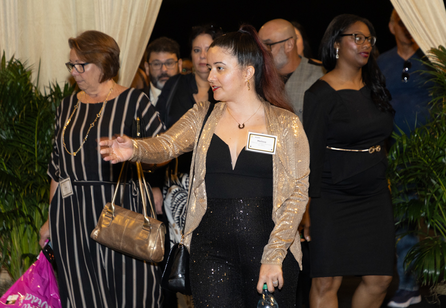 Woman in a gold sequin blazer leading a group at a formal event; she wears a name badge and black outfit, centered among attendees.