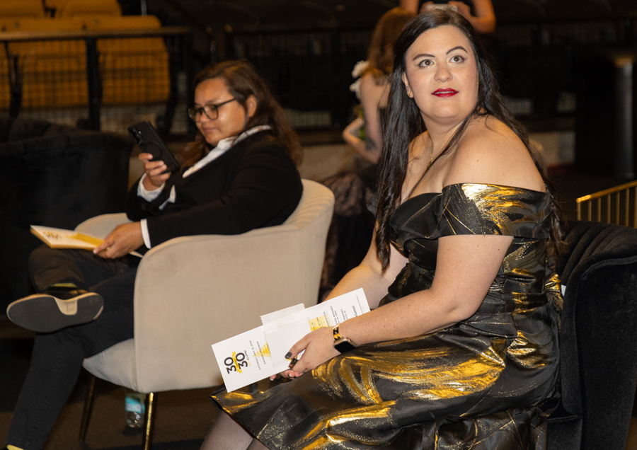 Woman in a metallic gold off-shoulder dress sits in a theater, holding event programs; another attendee uses a phone in the background.
