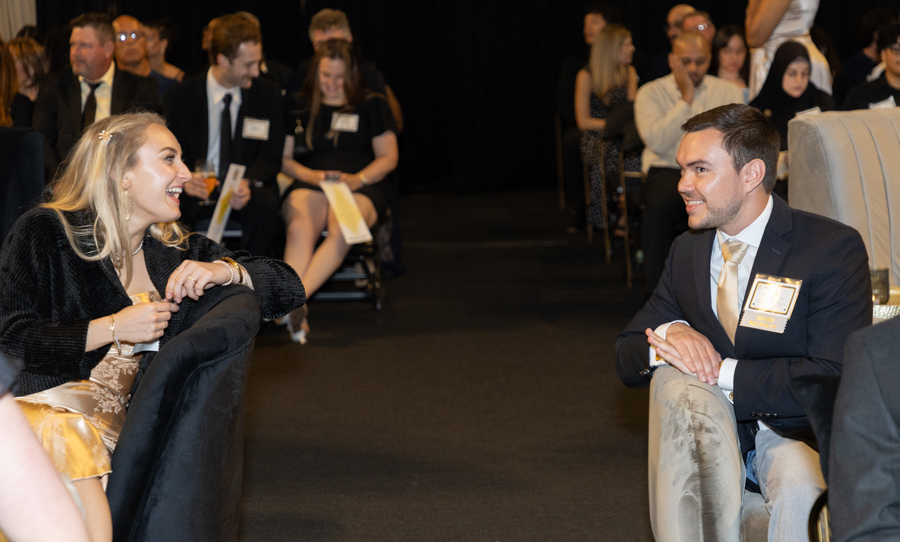 A woman in a gold dress and black jacket laughs while facing a man in a suit at a formal event, seated and chatting as other guests look on in the background.