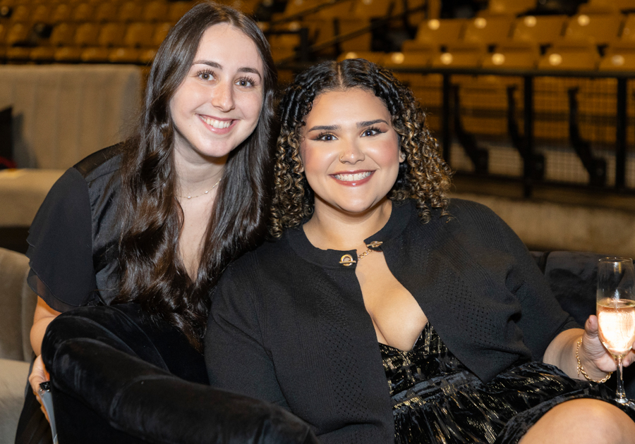 Two smiling women sit on a couch at an event, one holding a champagne flute in a dark venue backdrop.