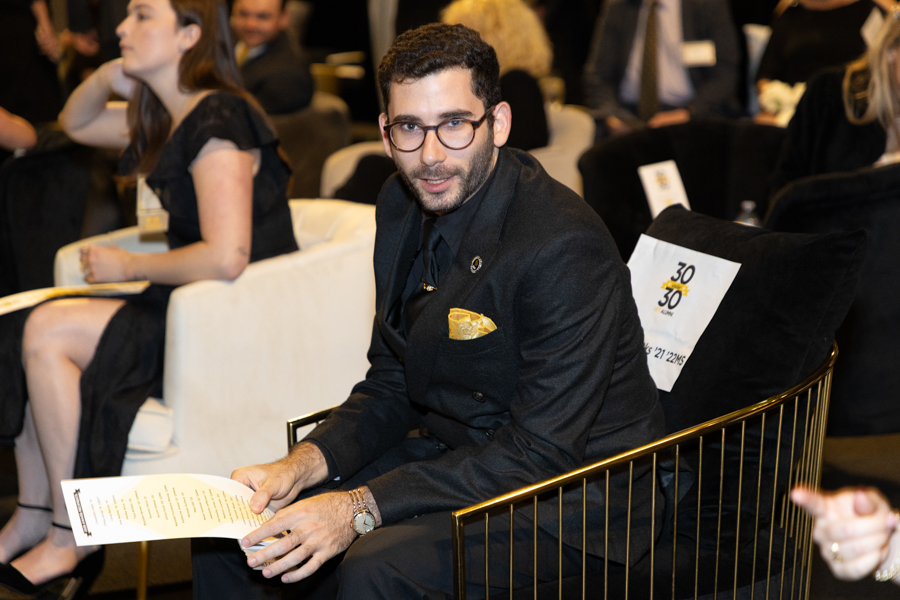 Man in a black suit and glasses sits at a formal event, reading a program booklet.