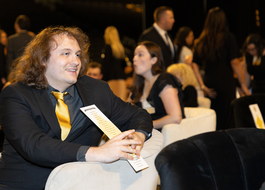 Man in a dark suit with a gold tie sits on a white sofa at a formal event, holding a program and smiling.