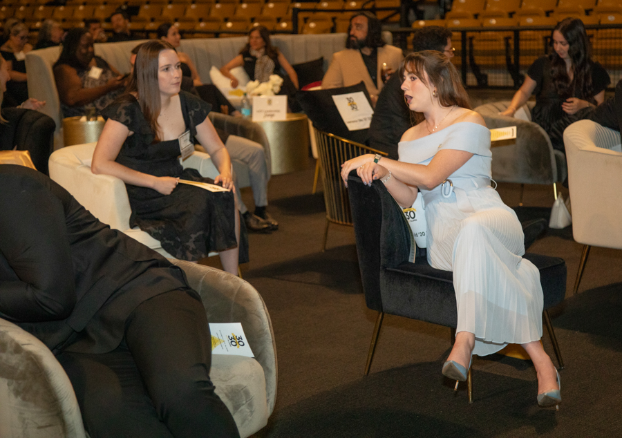 Two women seated in a lounge at a formal event, one in a black dress listening to the other in a light blue off-shoulder dress.
