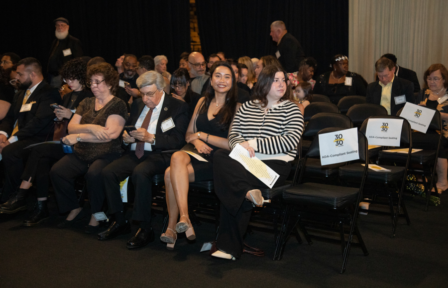 Group of diverse people seated in a conference hall, some looking at phones, front row smiling at the camera.