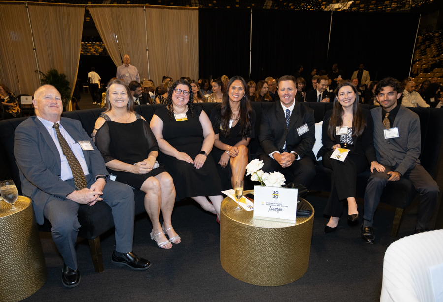 Seven attendees in formal attire sit on a black sofa at a gala, smiling for the camera.