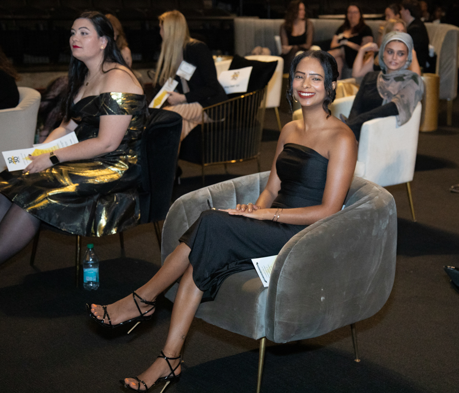Woman in a black strapless dress sits in a gray curved chair at a formal event, smiling at the camera, with others seated in the background.