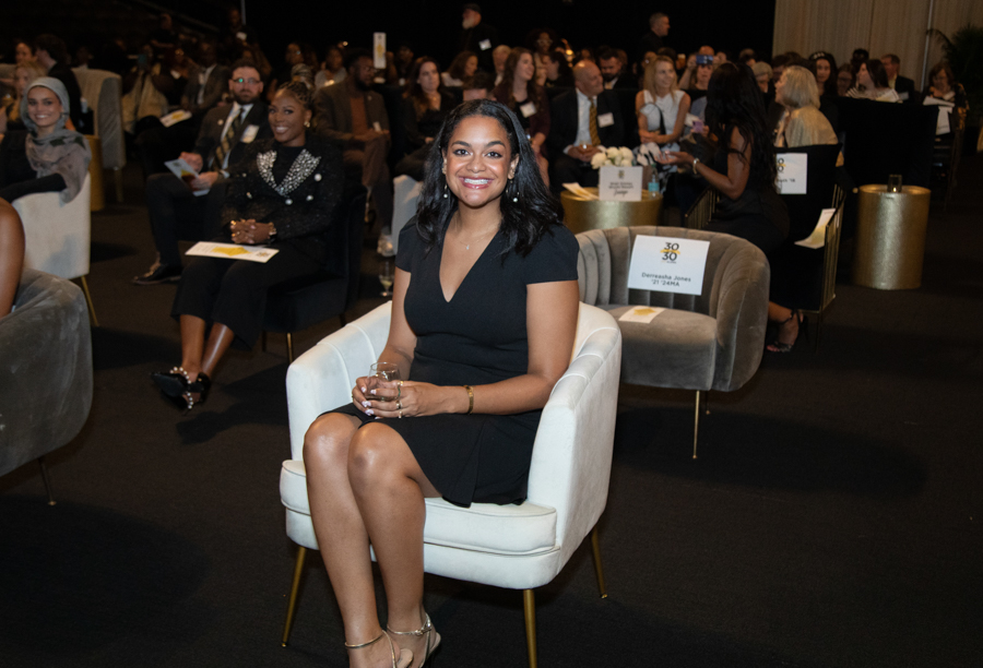 Smiling woman in a black dress seated in a white chair at a formal event with attendees in the background.