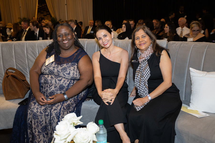 Three women in formal dresses sit on a gray sofa at a gala, smiling for the camera.