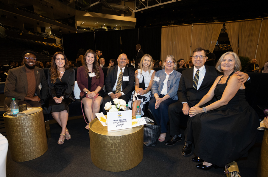 Nine adults in business attire sit together in a row at a formal event, posing for a group photo with smiles.